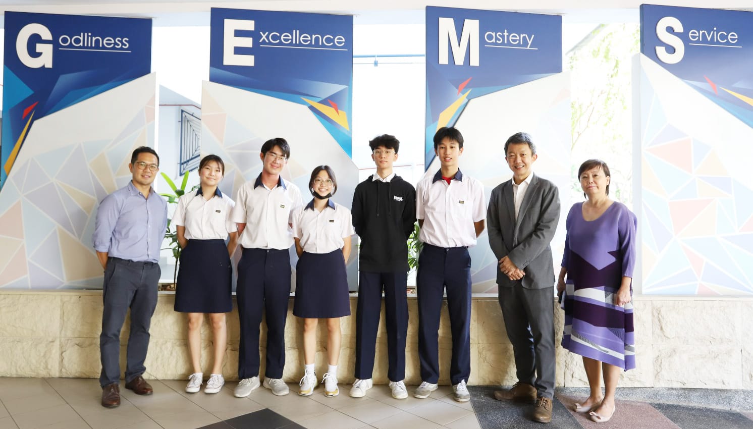 Eight people pose in front of banners reading Godliness, Excellence, Mastery, and Service at a school.
